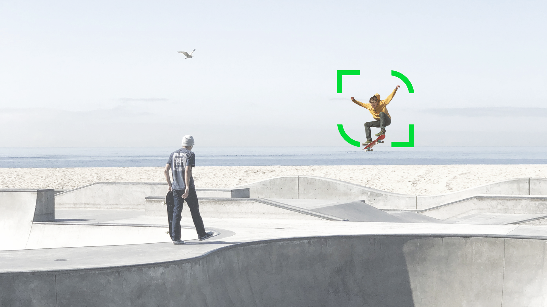 Two skaters in a concrete skate park, one jumping, the other holding his board. The sea and a seagull can be seen in the background.