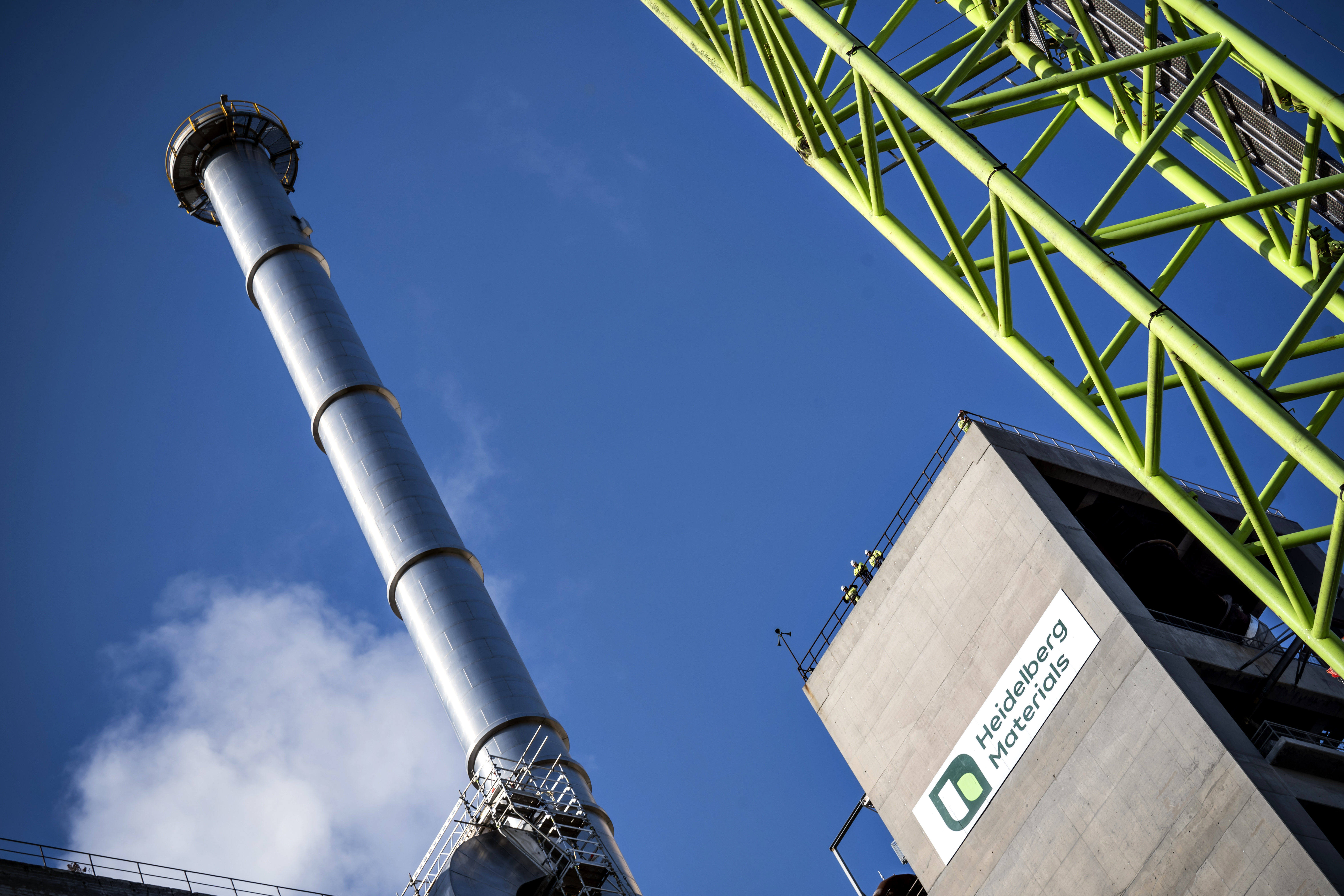 Industrial facility with a tall metal chimney emitting a small amount of white steam on the left, a green steel framework on the right, and a concrete building displaying a company logo and text in the bottom right; blue sky in the background.