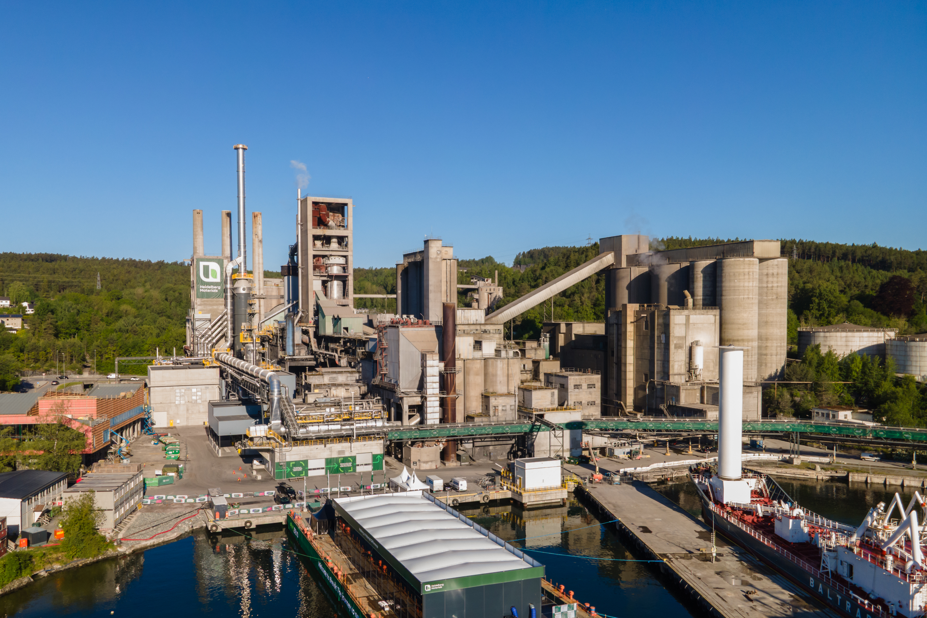 Brevik CCS plant: A large industrial cement plant with multiple tall chimneys, silos, and connected structures, surrounded by greenery and hilly terrain in the background. In the foreground, there is a water channel with docks, industrial equipment, and a white ship partially visible on the right. The setting is under a clear blue sky.