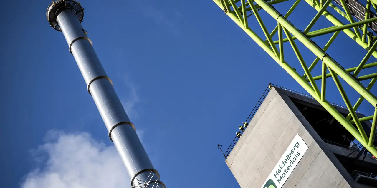Industrial facility with a tall metal chimney emitting a small amount of white steam on the left, a green steel framework on the right, and a concrete building displaying a company logo and text in the bottom right; blue sky in the background.