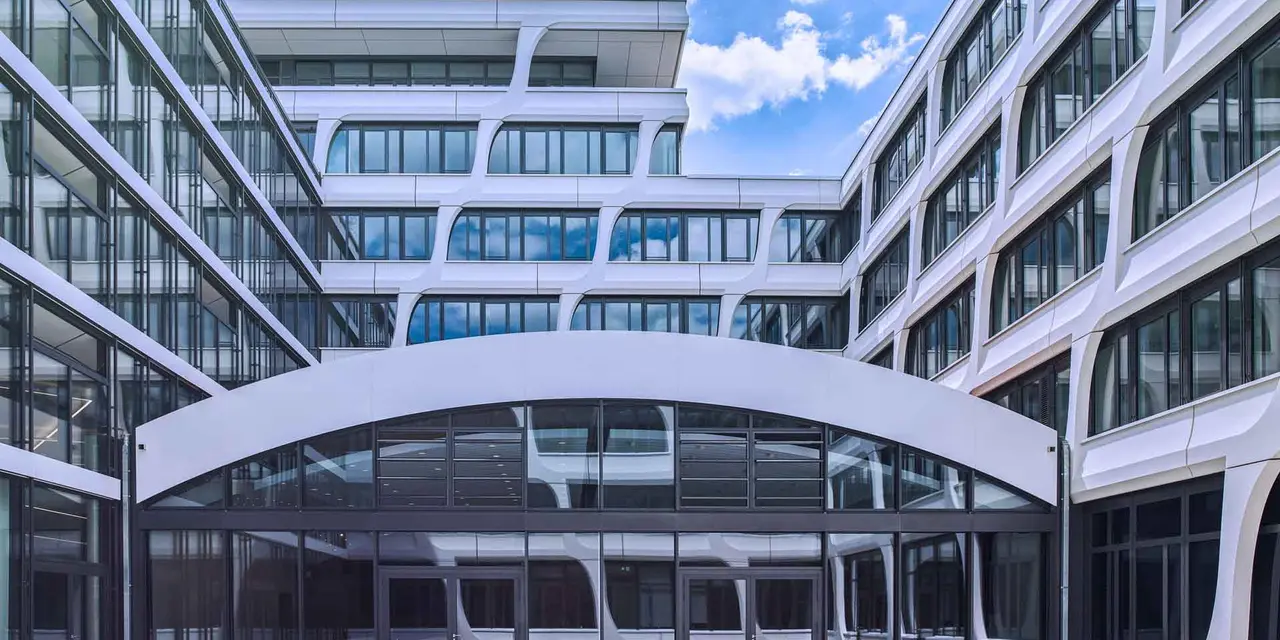 A modern, white office building with a curved glass atrium and a central courtyard. The building is surrounded by other office buildings and has a blue sky in the background.