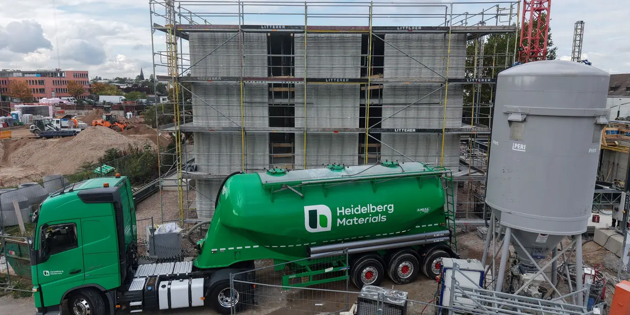 Camion verde con logo Heidelberg Materials in un cantiere edile con impalcature e attrezzature industriali.