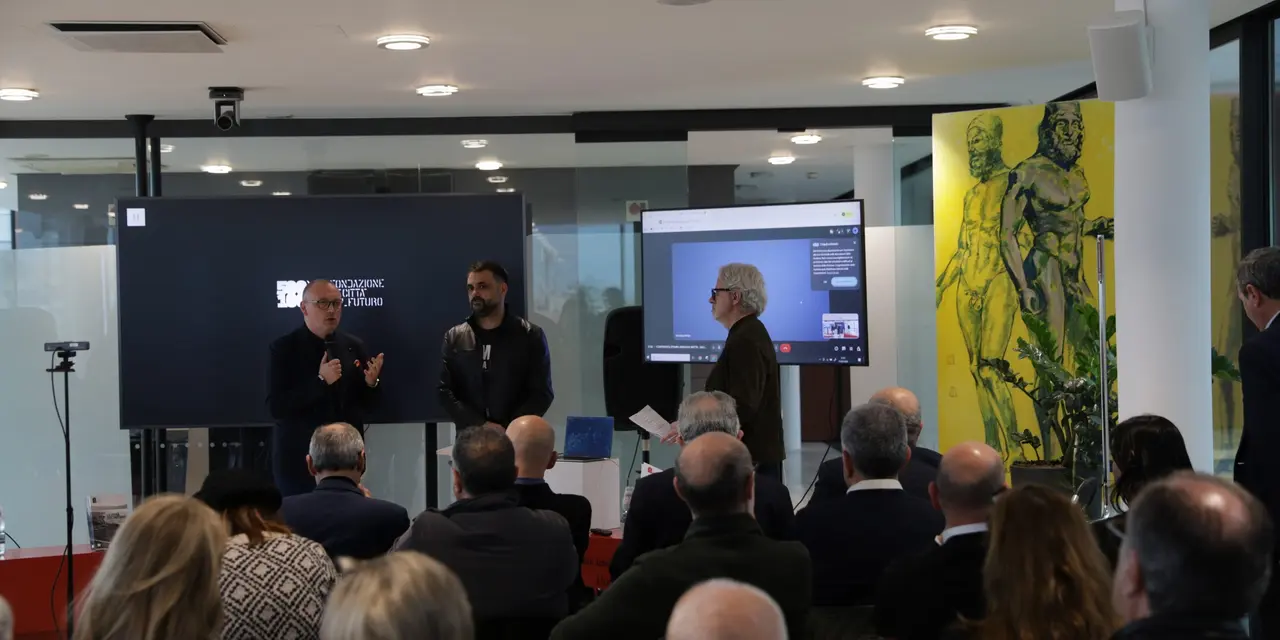 Persone riunite in una sala per una presentazione aziendale, con schermo e opere d'arte visibili sullo sfondo.