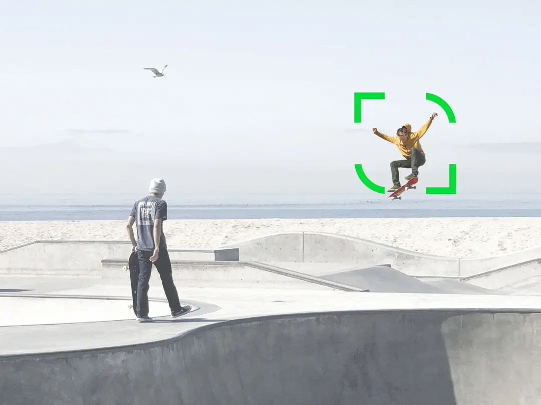 Two skaters in a concrete skate park, one jumping, the other holding his board. The sea and a seagull can be seen in the background.