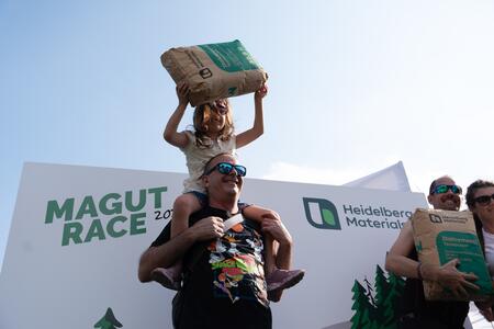 Una bambina sollevata sulle spalle di un uomo tiene un sacco di materiale da costruzione sopra la testa durante un evento Heidelberg Materials.