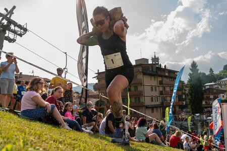 Una persona partecipa a una gara trasportando sacchi di cemento su una collina, con edifici e pubblico sullo sfondo.