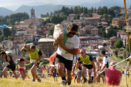 Persone che corrono su una collina trasportando sacchi di cemento, con un paesaggio urbano sullo sfondo.
