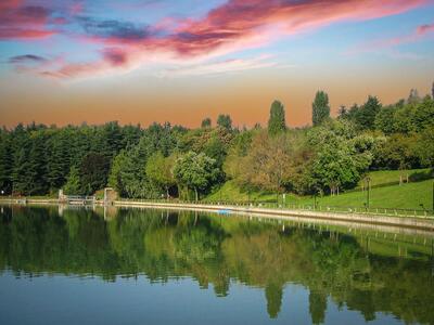 Un parco con un lago riflettente, circondato da alberi e un cielo al tramonto.